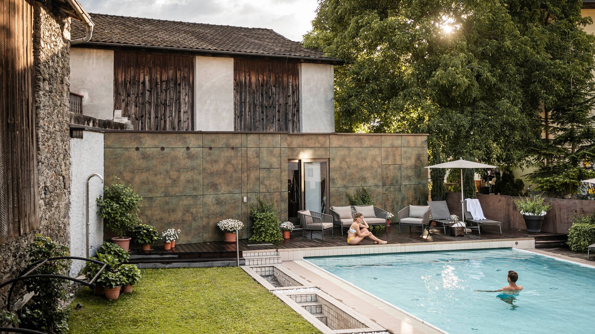 Una piscina circondata da un giardino curato, con lettini e ombrelloni. Una persona è seduta a bordo piscina, mentre un'altra nuota. Sullo sfondo, una struttura con pareti in legno e verde. Il sole splende tra gli alberi.
