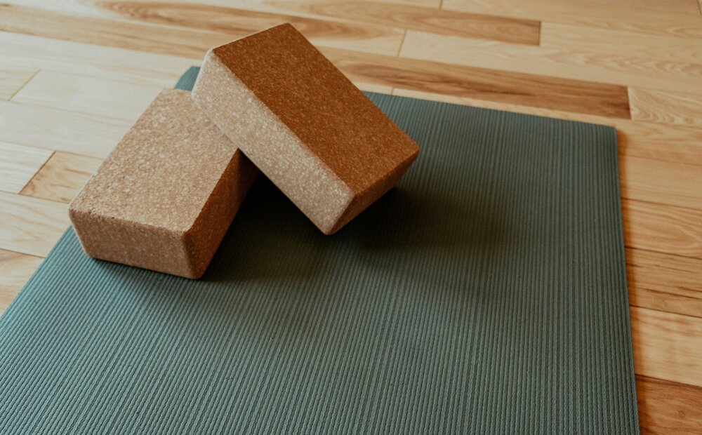 Two cork yoga blocks are positioned on a textured green yoga mat, set against a backdrop of light wooden flooring. The scene conveys a calm and inviting atmosphere, ideal for yoga practice.