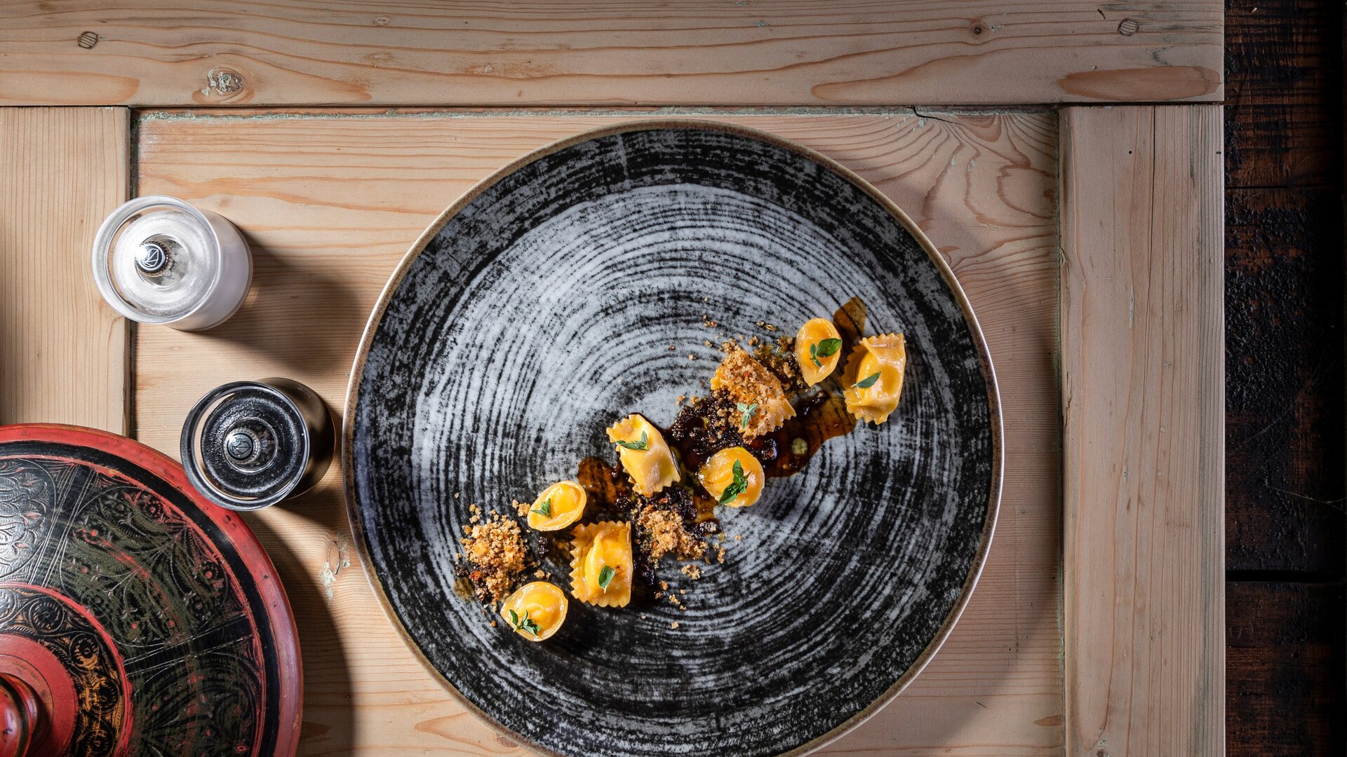 A beautifully arranged plate of pasta sits on a textured black dish, garnished with herbs and breadcrumbs. The setting includes a wooden table with a decorative lid and salt and pepper shakers nearby, creating an inviting dining atmosphere.