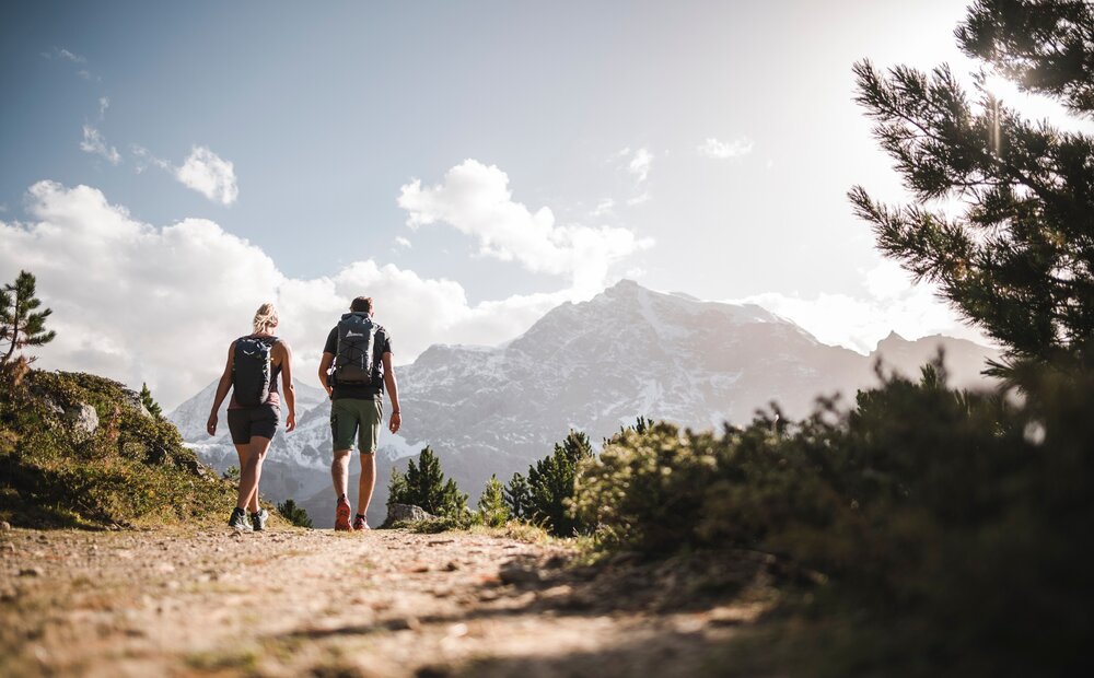 Two hikers walk along a dirt path surrounded by greenery, with a backdrop of majestic mountains under a partly cloudy sky. The sun casts a warm glow, enhancing the natural beauty of the landscape.