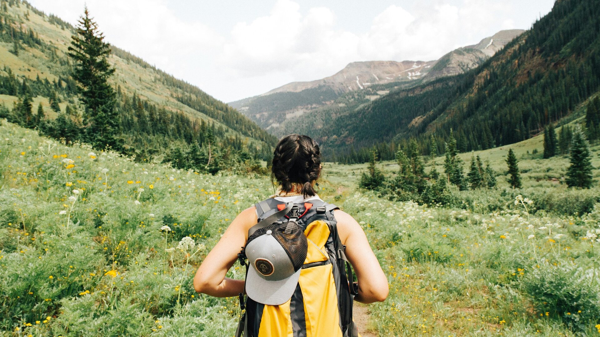 A person with braided hair is walking along a trail in a lush green valley, surrounded by wildflowers and tall trees. The backdrop features mountains under a partly cloudy sky. The individual is wearing a backpack and a cap, facing away from the camera.