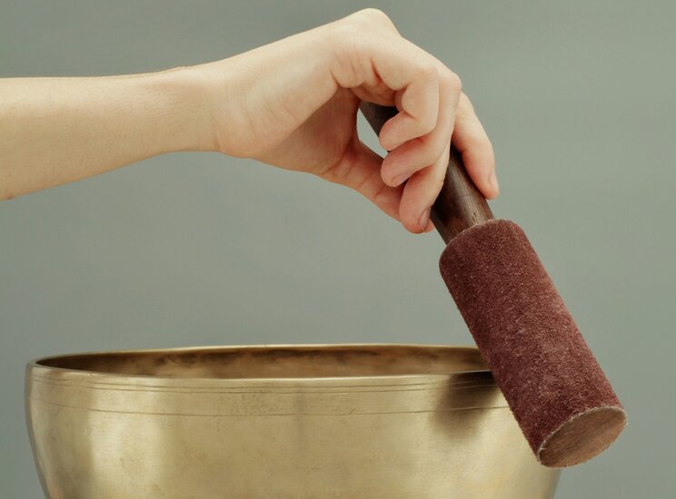 Una mano tiene un bastone rivestito di feltro sopra una ciotola di metallo dorato. Lo sfondo è grigio chiaro, creando un'atmosfera calma e meditativa.