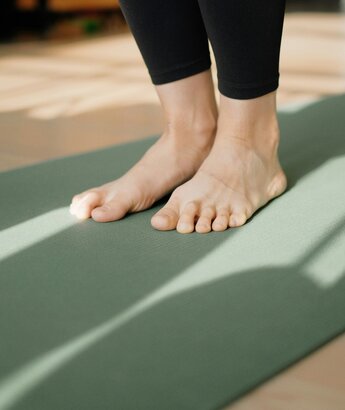 Due piedi nudi poggiano su un tappetino da yoga verde, con luce naturale che filtra creando ombre sul pavimento. L'atmosfera è calma e rilassante, ideale per la pratica dello yoga.