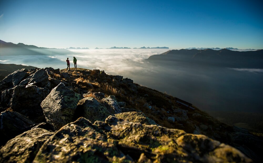 Two hikers stand on a rocky mountain peak, overlooking a sea of clouds below. The sun casts a warm glow, illuminating the landscape and distant mountain ranges under a clear blue sky.