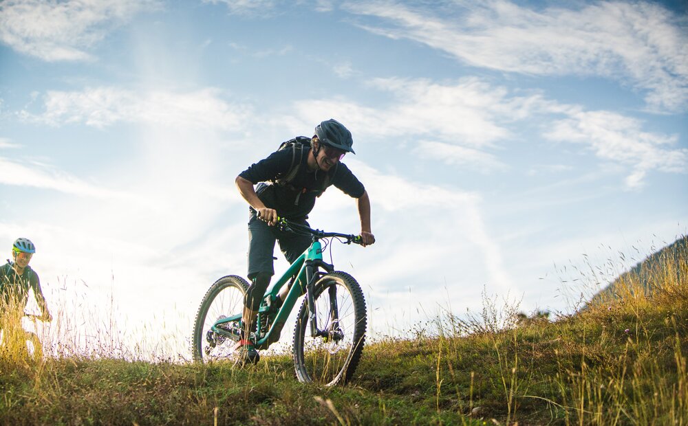 A mountain biker climbs a grassy hill, focused on navigating the terrain. Another cyclist is visible in the background, enjoying the outdoor setting under a bright sky with scattered clouds. The scene captures the thrill of mountain biking in nature.
