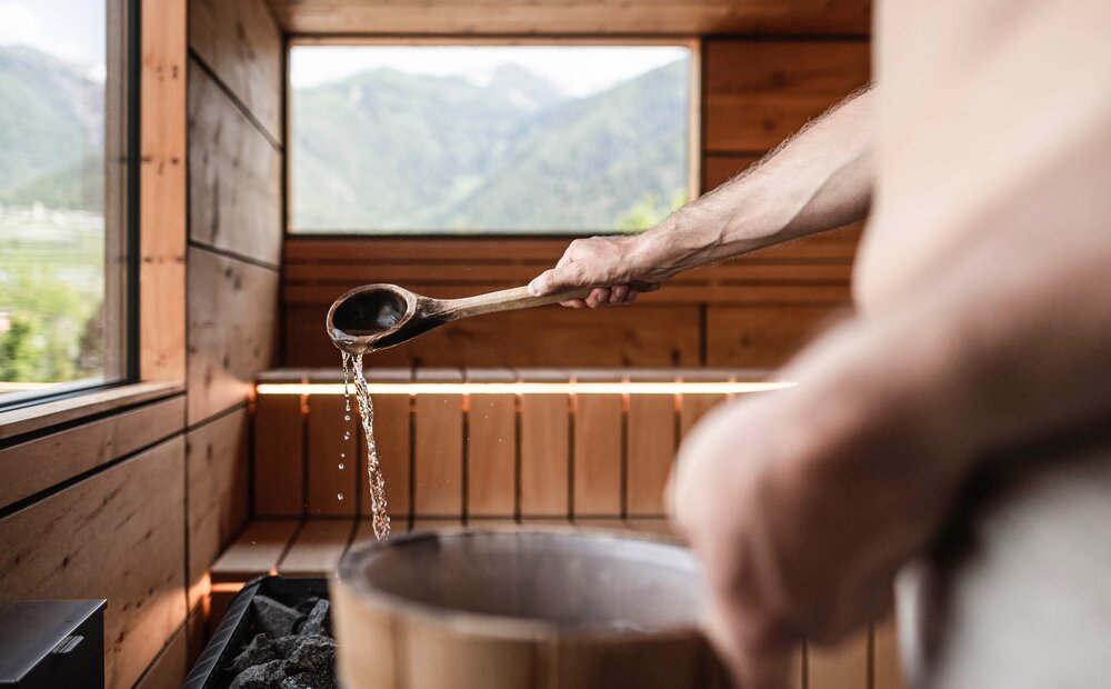 Interno di una sauna in legno con vista su montagne verdi. Un uomo versa acqua da un mestolo di legno su delle pietre calde. L'atmosfera è rilassante e naturale, con luce che filtra attraverso le finestre.
