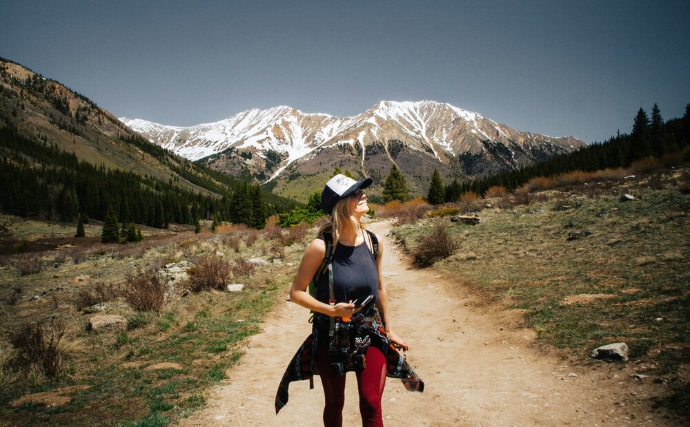 A woman walks along a dirt path in a mountainous landscape, wearing a cap and a backpack. She smiles while looking up at the snow-capped peaks in the background, surrounded by green trees and shrubs. The sky is clear and blue.