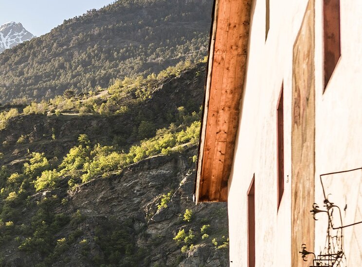 Offerta wellness Val Venosta – Hotel Matill a Laces Una parete di una casa con finestre rosse si affaccia su una montagna verdeggiante, con rocce e alberi. Sullo sfondo, si vedono cime innevate sotto un cielo sereno. La luce del sole illumina la scena, creando un'atmosfera tranquilla.