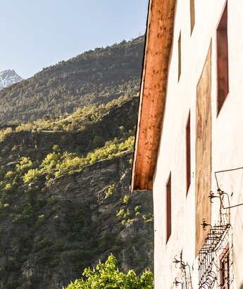 Offerta wellness Val Venosta – Hotel Matill a Laces Una parete di una casa con finestre rosse si affaccia su una montagna verdeggiante, con rocce e alberi. Sullo sfondo, si vedono cime innevate sotto un cielo sereno. La luce del sole illumina la scena, creando un'atmosfera tranquilla.