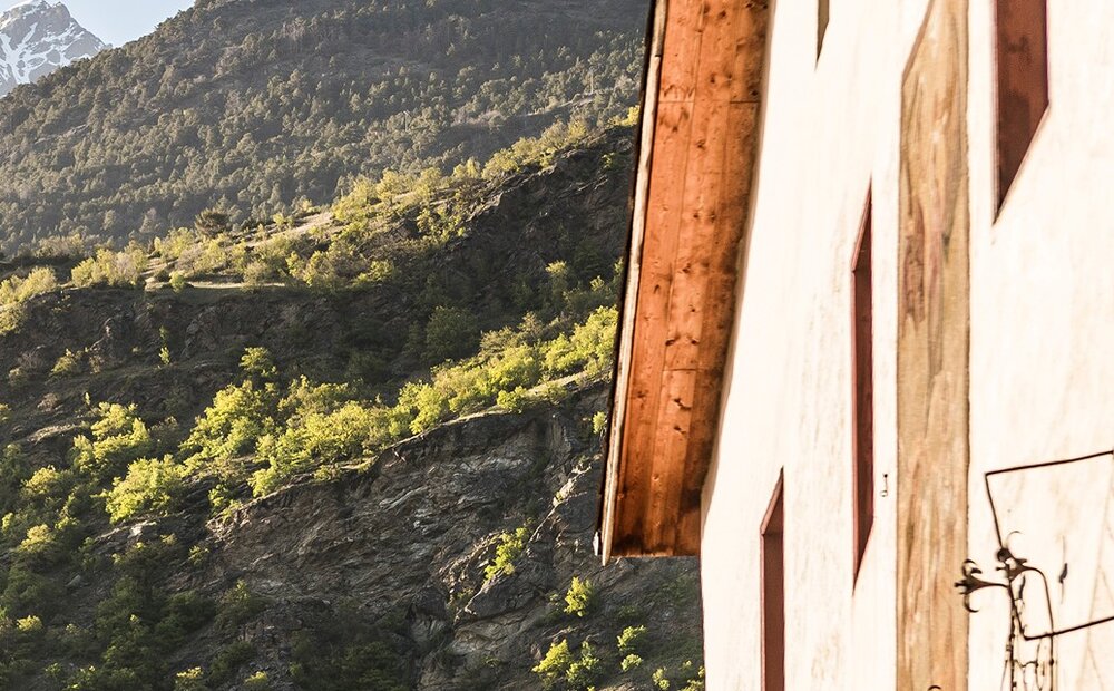 Offerta wellness Val Venosta – Hotel Matill a Laces Una parete di una casa con finestre rosse si affaccia su una montagna verdeggiante, con rocce e alberi. Sullo sfondo, si vedono cime innevate sotto un cielo sereno. La luce del sole illumina la scena, creando un'atmosfera tranquilla.