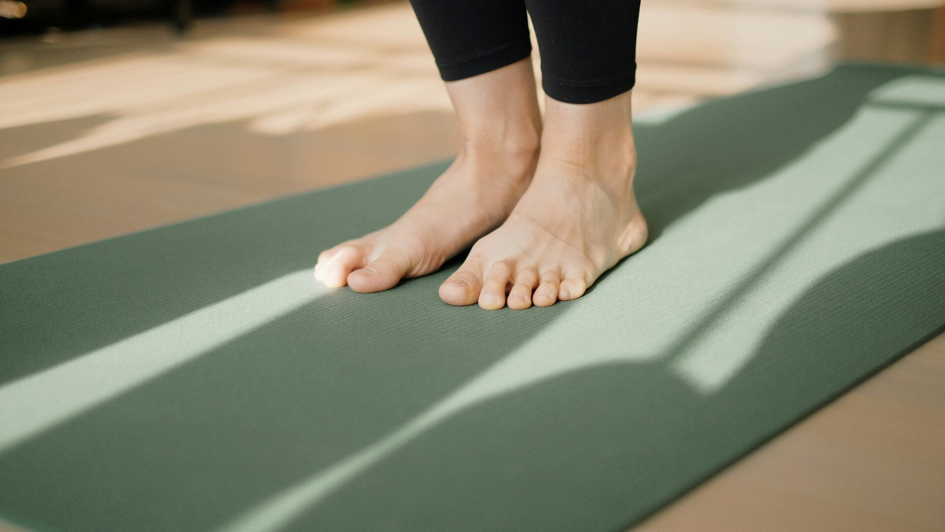 A close-up of bare feet standing on a green yoga mat, with sunlight casting soft shadows on the mat and floor. The background is softly blurred, suggesting a calm indoor environment.