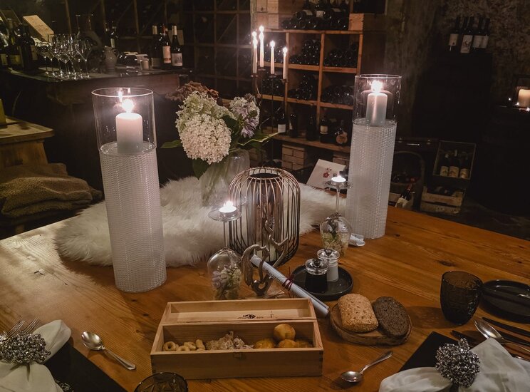 Tavolo elegantemente apparecchiato in una cantina, con candele accese, un vaso di fiori bianchi e una selezione di cibi su un vassoio di legno. Sullo sfondo, scaffali di vino e bicchieri. Atmosfera intima e accogliente.