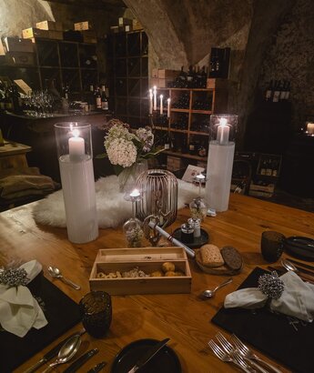 Ein elegant gedeckter Tisch in einem gemütlichen Weinkeller. Auf dem Tisch stehen Kerzen, eine Blumenvase mit weißen Blumen und eine Holzkiste mit Snacks. Im Hintergrund sind Weinregale und Gläser sichtbar, die eine einladende Atmosphäre schaffen.