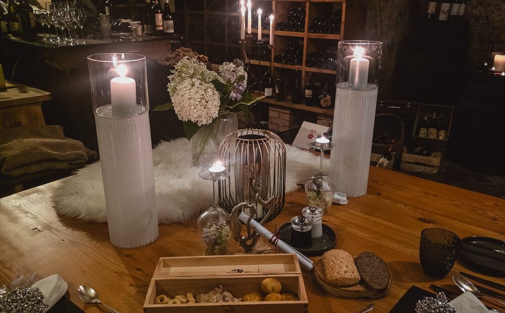 A cozy dining table set with elegant tableware, white napkins, and decorative elements. The table features a wooden box with snacks, candles, and a vase of flowers. In the background, wine bottles are displayed on shelves, creating a warm, intimate atmosphere.