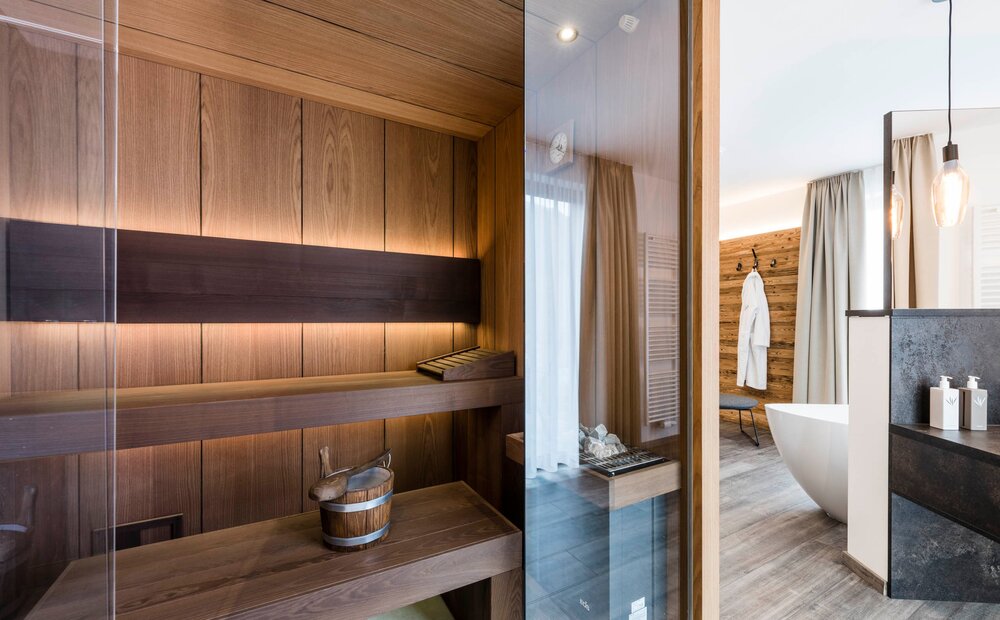 Interno di una sauna in legno con panchine e un secchio in legno. Si intravede un bagno moderno con una vasca bianca, un appendiabiti con un accappatoio e pareti in legno. Illuminazione calda e atmosfera rilassante.