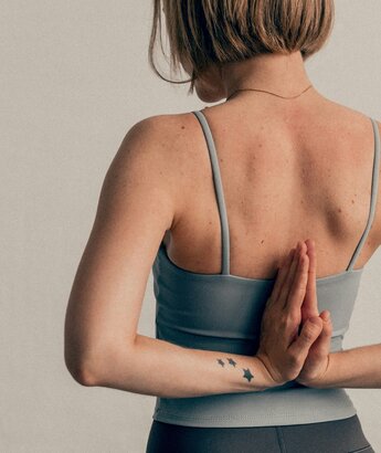 A person with short hair is seen from behind, wearing a light blue tank top. Their hands are clasped together in a prayer position at the back, and a small tattoo of three stars is visible on their forearm. The background is a soft, neutral color.